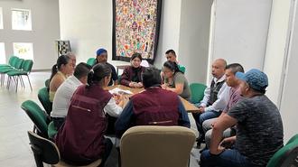 Group of people seated around a round table in a bright room, a quilt hanging on the wall.