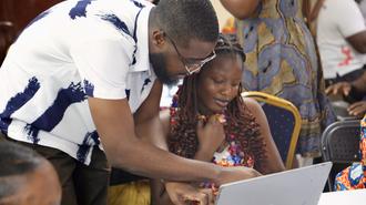 Photograph of a man helping a girl with a laptop at a table outdoors.