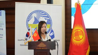 Woman speaks at a podium during a formal event, with the Kyrgyzstan flag and banners behind.