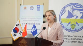 Photograph: woman in pale pink blazer speaks at a podium with flags.
