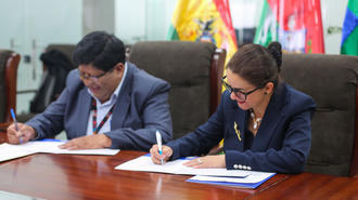 Two professionals signing documents at a conference table, colorful flags in the background.