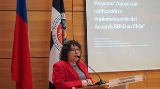 Photograph of a woman in a red blazer speaking at a podium with flags and a projection screen.