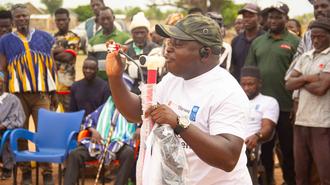 Man in white shirt and camouflage cap speaks into a microphone to an outdoor crowd.