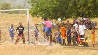 Photograph of a dusty soccer field with players near the goal and a crowd watching.