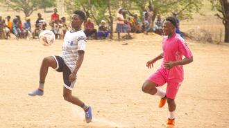 Photo of two players chasing a ball on a dusty field; a girl in pink and a boy in white.