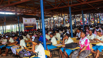 Photograph of a crowded classroom with students at long desks under a wooden ceiling and blue posts