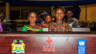 Group of women seated behind a banner featuring a crest and the UNDP logo.