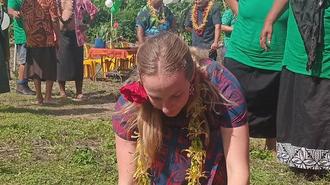 Girl kneeling to plant in dirt at an outdoor community event; volunteers in green shirts nearby.
