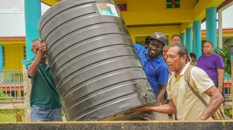 Men help lift a large gray water tank beside a colorful yellow-and-teal building.