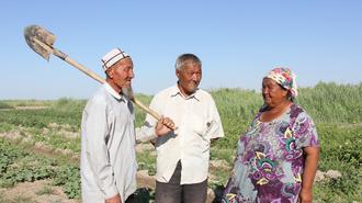 Three people stand in a sunlit field, conversing; one holds a long-handled tool with a curved blade.
