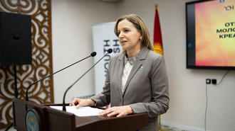 Photograph of a woman in a gray suit at a podium giving a speech in a conference room.