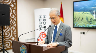 Senior man in a suit speaks at a podium with banners, flags, and a screen behind.