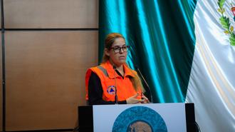 Speaker in an orange safety vest at a podium, with a teal curtain backdrop.