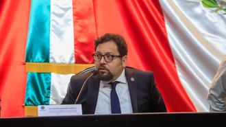 Man in suit and glasses speaks at a panel, with red, white, and blue flags behind.