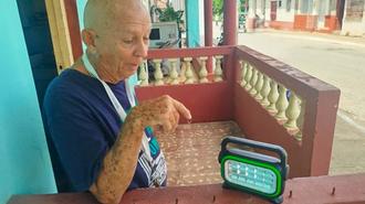 Older man in a blue shirt sits on a porch, holding a water bottle beside a green digital clock.