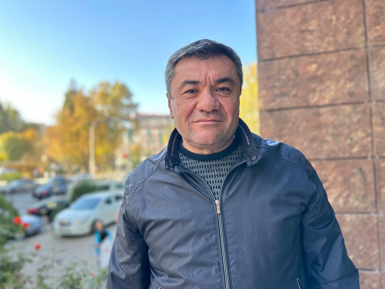 A man stands outdoors with a slight smile, facing the camera. He is wearing a dark zippered jacket over a sweater with a geometric pattern. The background is softly blurred, showing a sunny, urban setting with trees in autumn colors and parked cars.