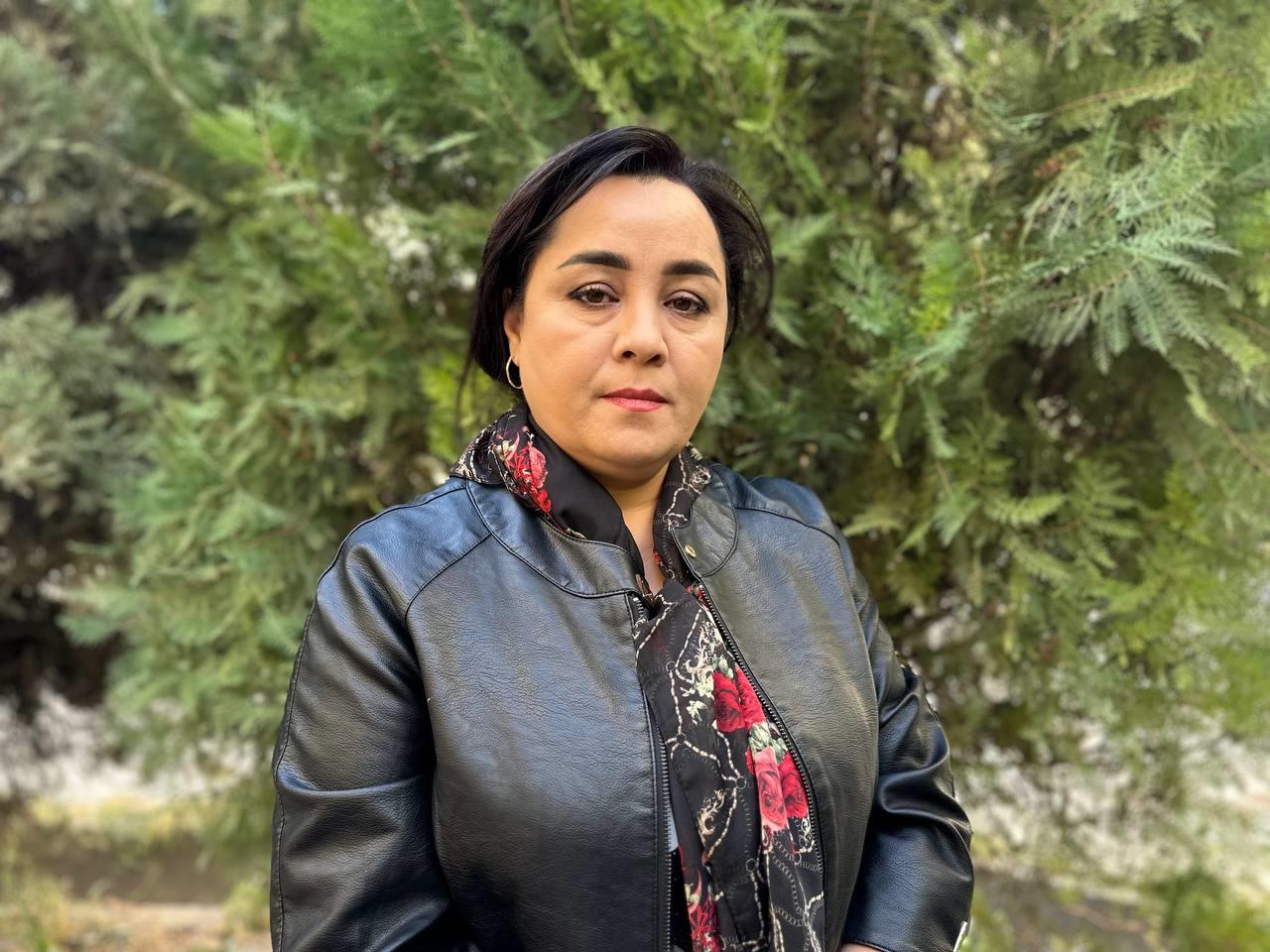 A woman with medium-length hair, wearing a black leather jacket and a scarf with red floral patterns, stands in front of evergreen trees.