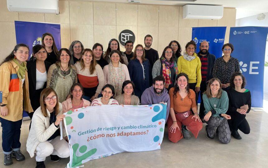 Foto grupal de docentes y referentes de Clubes de ciencia junto a talleristas. Cuadro de las personas lleva una cartel con la inscripción: "Gestión de riesgo y cambio climático, ¿cómo adaptarnos?"