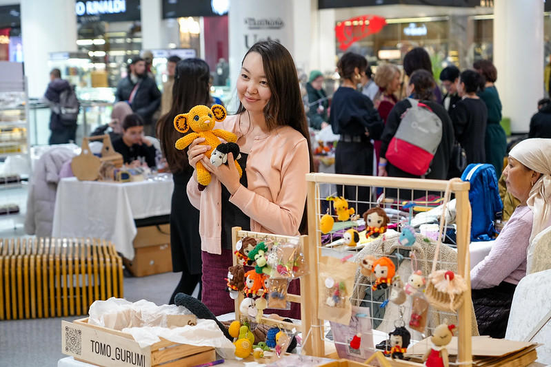 A young girl with long hair holding a hand-made toy near the table with similar toys during the exhibition. 