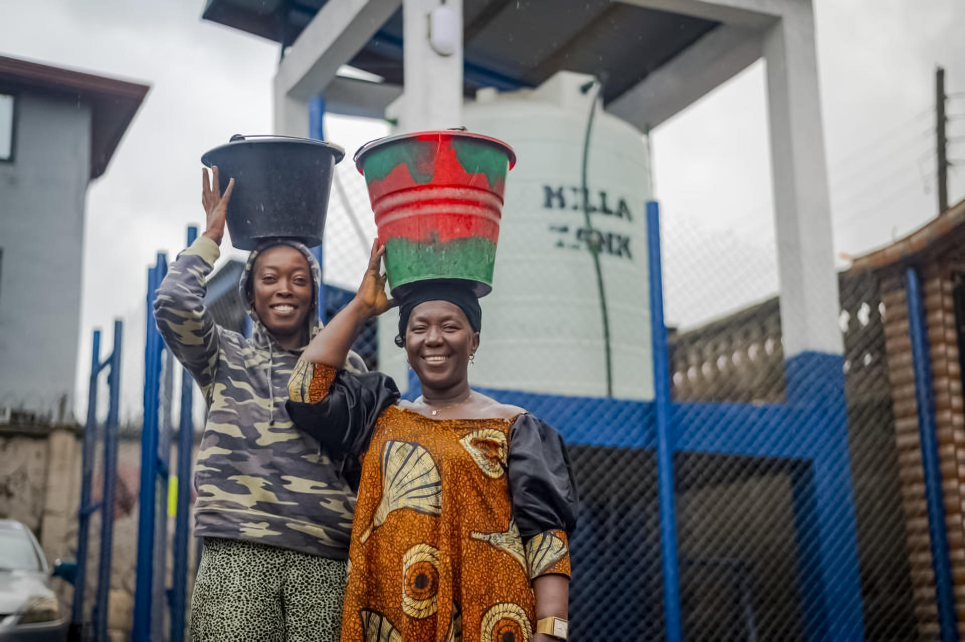 two women with buckets on their heads standing in front of water tank