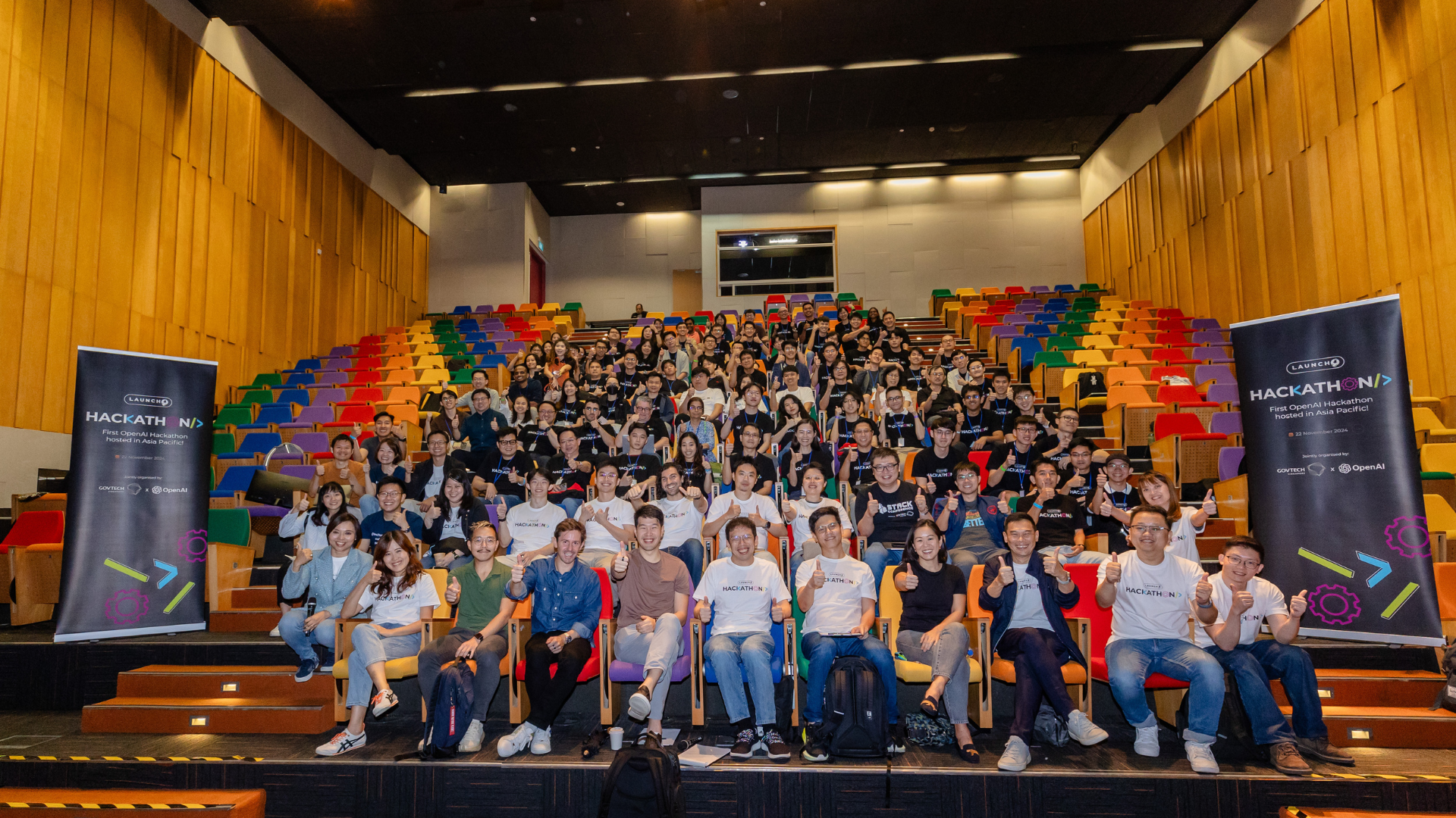 A group of people in an auditorium with two pull up banners on both sides signifying that a Hackathon was held.
