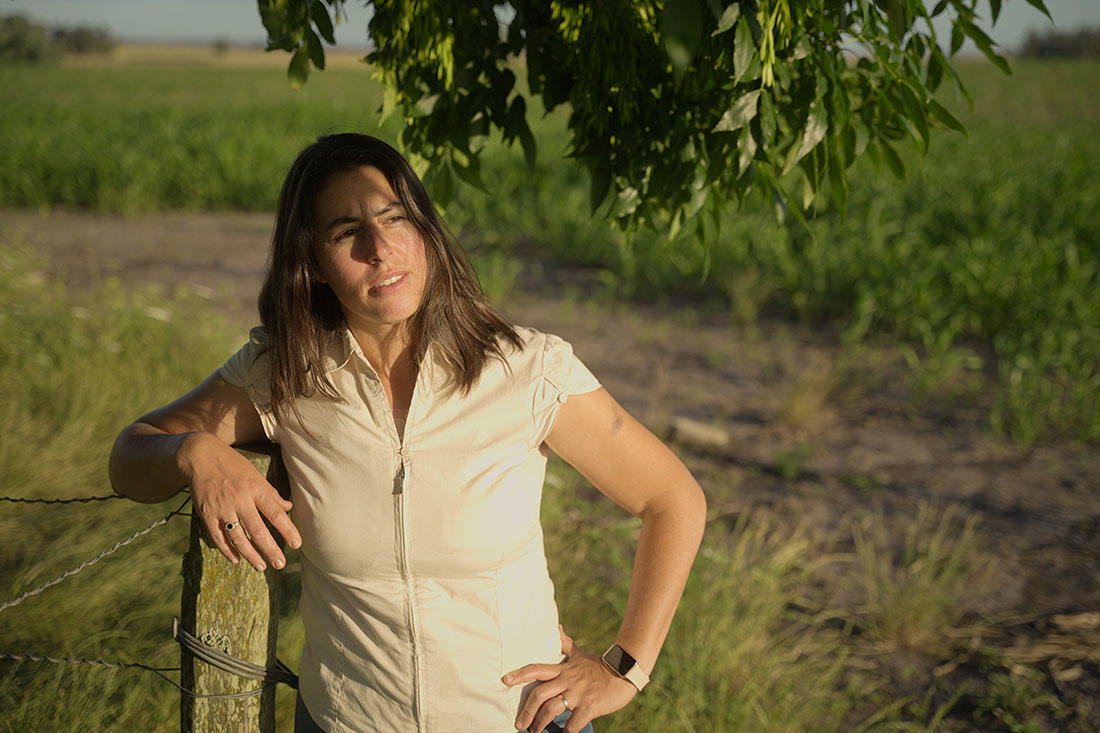 Karen, una mujer rural en Lavalleja, está apoyada en un poste de madera junto a un alambrado, rodeada de un campo verde bajo la luz del sol. Lleva una camisa beige de manga corta y un reloj inteligente en la muñeca. Su mirada se dirige hacia un punto en la distancia, con una expresión pensativa. Al fondo, se aprecian cultivos y el horizonte despejado, reflejando un entorno rural sereno.
