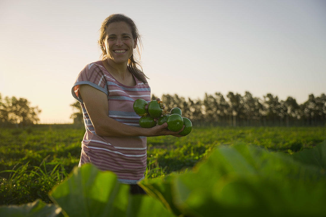 Rosina, una mujer rural en San José, está de pie en un galpón de trabajo con una sonrisa cálida. Lleva una camiseta de rayas en tonos rosados y grises. Detrás de ella, se observan cajas con manzanas y maquinaria agrícola, como montacargas, en un ambiente laboral rural. La luz natural ilumina la escena, destacando el esfuerzo y dedicación en su entorno de trabajo.