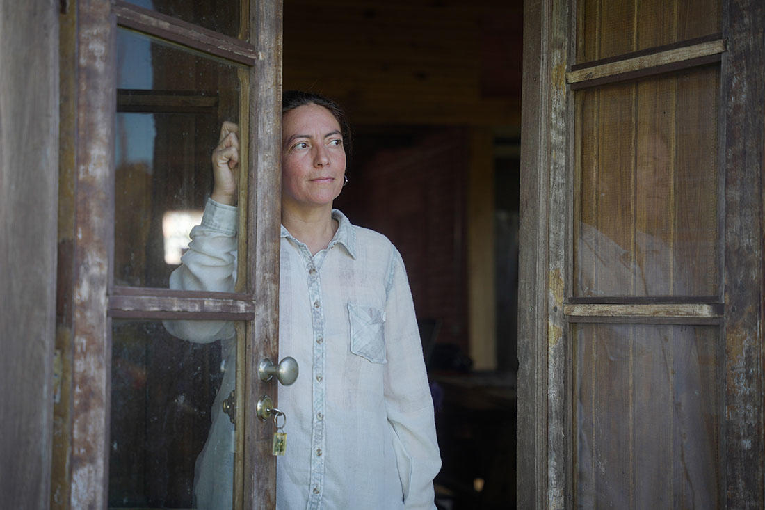Adriana, una mujer rural, aparece en el marco de una puerta de madera antigua con cristales. Lleva una camisa blanca y tiene una expresión reflexiva mientras mira hacia el exterior. En el interior, se distingue un espacio rústico con paredes de madera y ladrillo. La puerta tiene una cerradura con llave colgando, añadiendo un toque de autenticidad al entorno rural.