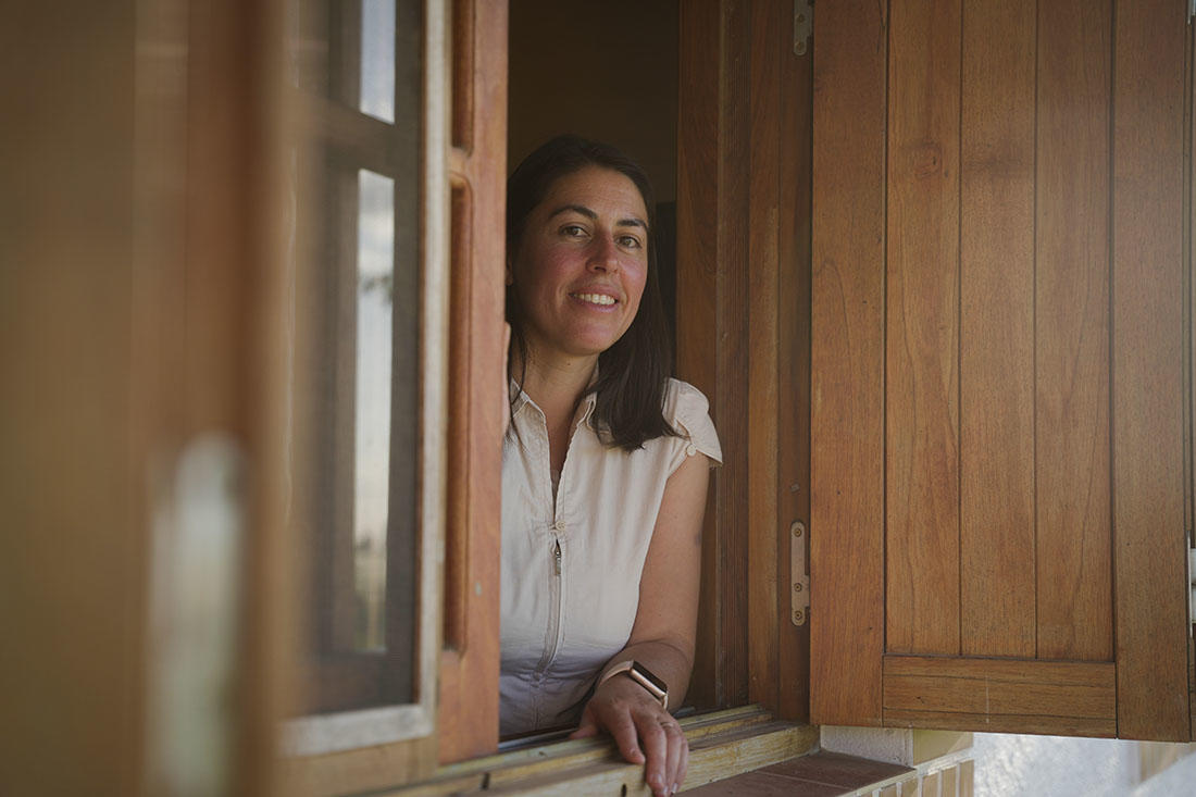 Karen, una mujer rural en Lavalleja, está apoyada en un poste de madera junto a un alambrado, rodeada de un campo verde bajo la luz del sol. Lleva una camisa beige de manga corta y un reloj inteligente en la muñeca. Su mirada se dirige hacia un punto en la distancia, con una expresión pensativa. Al fondo, se aprecian cultivos y el horizonte despejado, reflejando un entorno rural sereno.