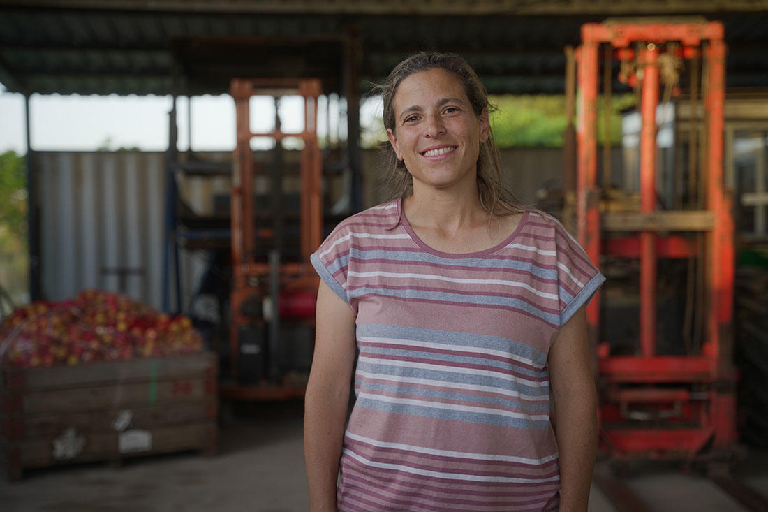Rosina, una mujer rural en San José, está de pie en un galpón de trabajo con una sonrisa cálida. Lleva una camiseta de rayas en tonos rosados y grises. Detrás de ella, se observan cajas con manzanas y maquinaria agrícola, como montacargas, en un ambiente laboral rural. La luz natural ilumina la escena, destacando el esfuerzo y dedicación en su entorno de trabajo.