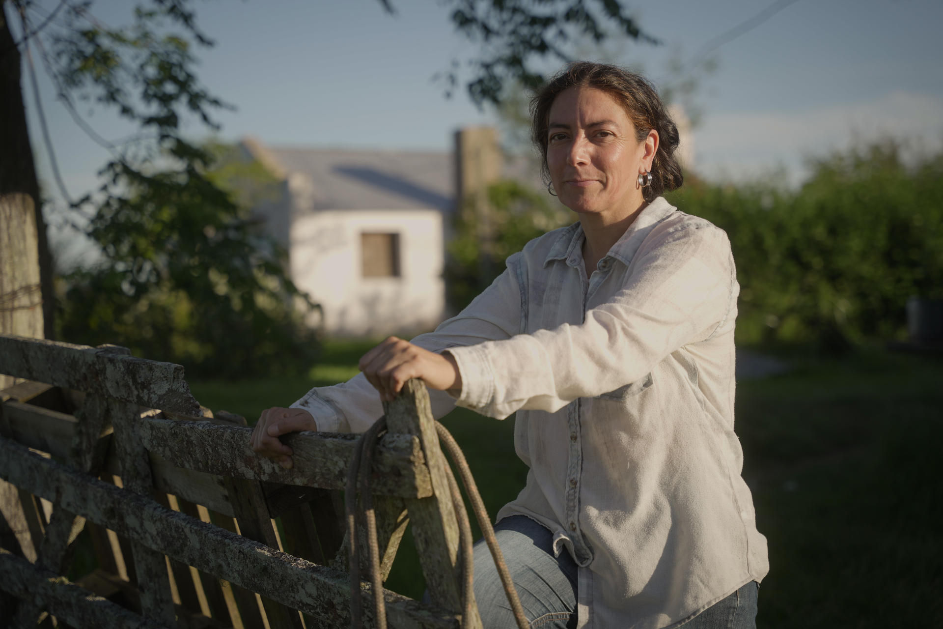 Adriana, una mujer rural en Rivera, aparece al aire libre apoyada en una cerca de madera desgastada, con una cuerda enrollada colgando. Lleva una camisa blanca y jeans, con una expresión serena. Al fondo, se observa una casa blanca parcialmente oculta por árboles y un cielo despejado. La escena transmite tranquilidad en un entorno rural.