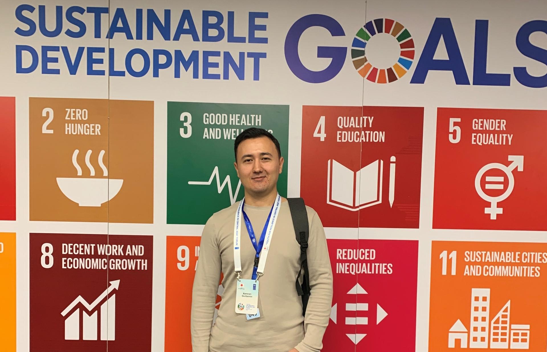 A young man stands in front of a colorful wall displaying Sustainable Development Goals.