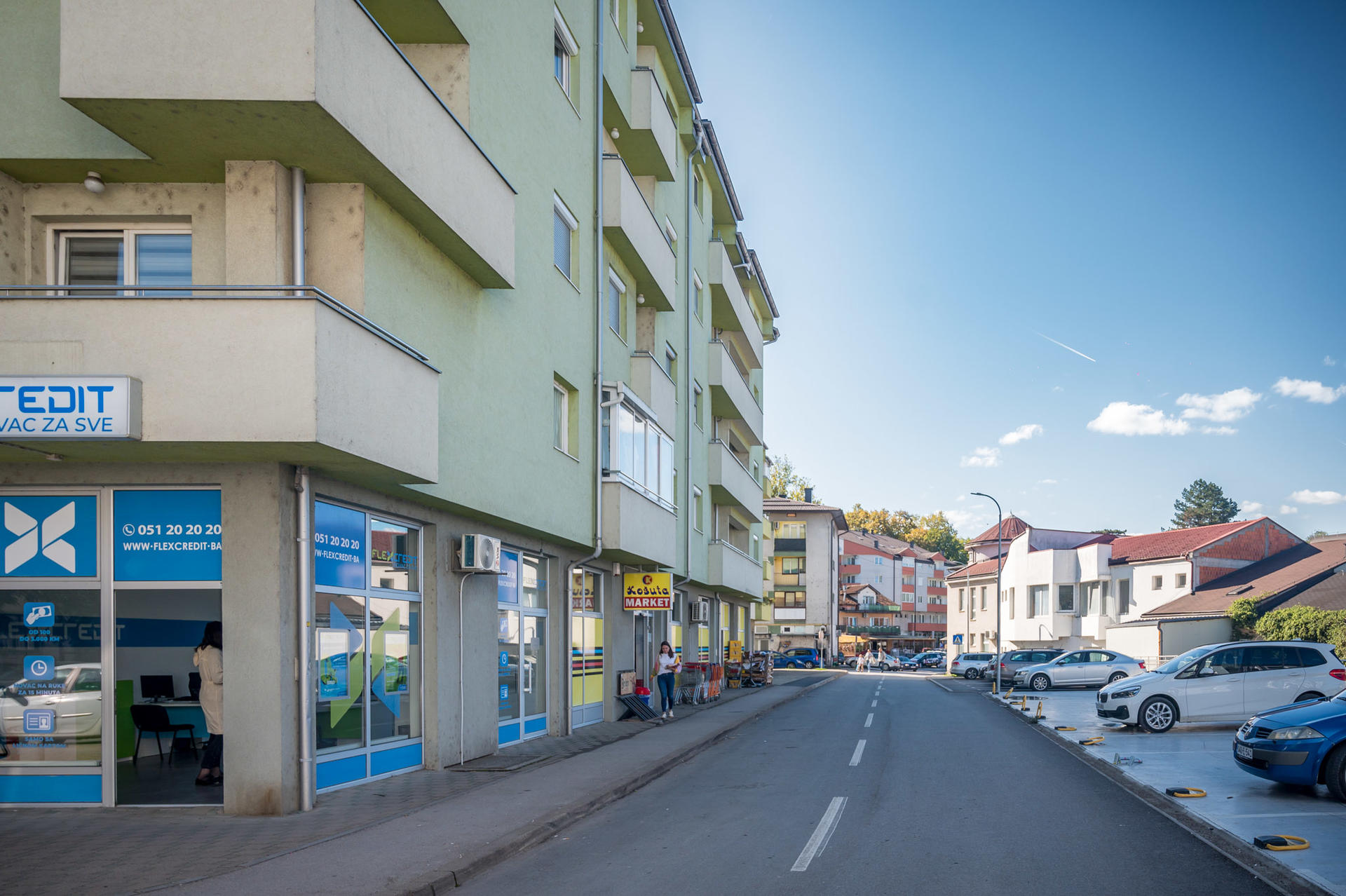 Street view featuring modern buildings, parked cars, and a clear blue sky.