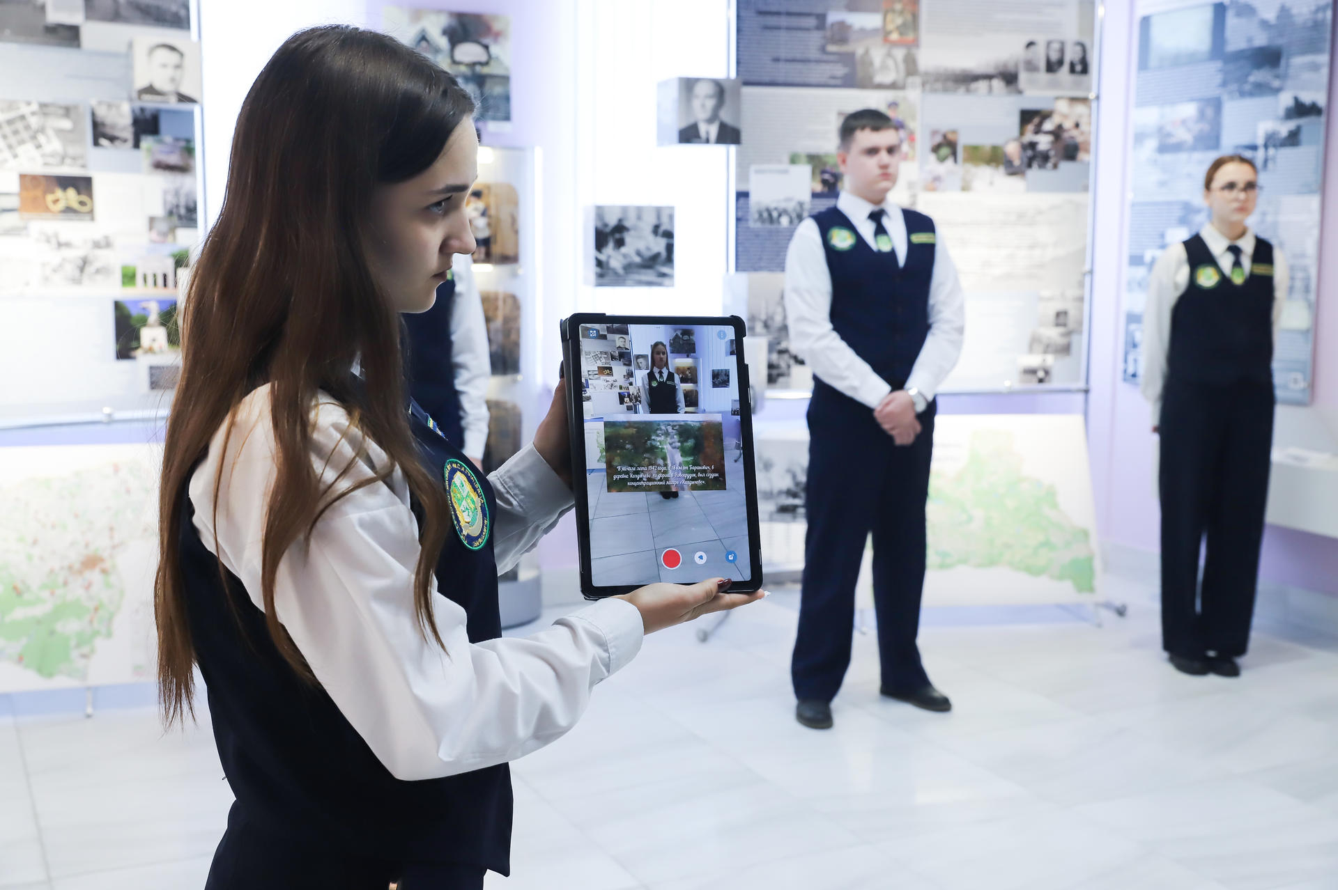A woman in a vest holds a tablet while two people stand in the background.