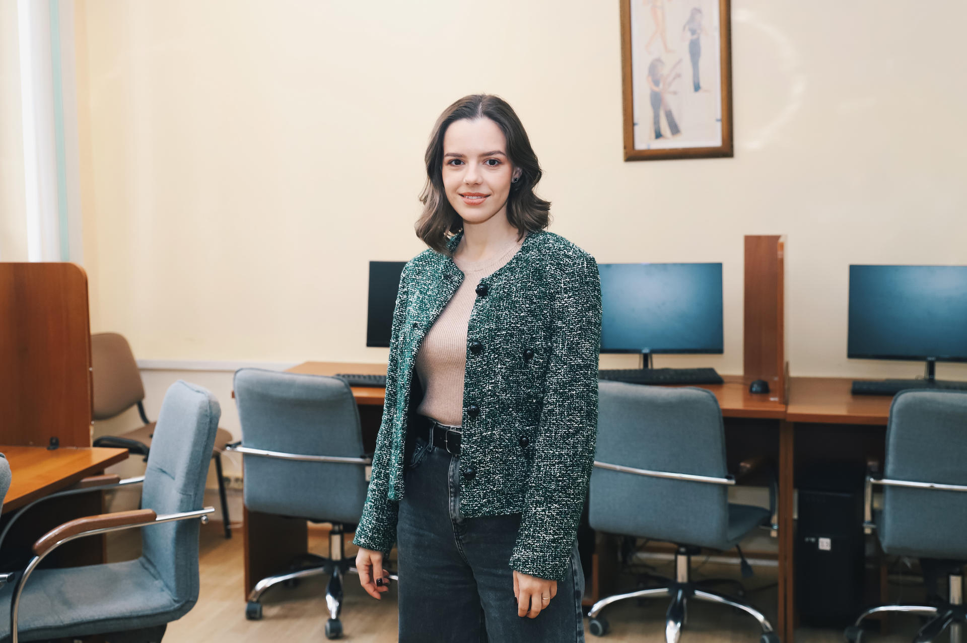 Young woman in a green sweater stands in a room with computer desks and chairs.