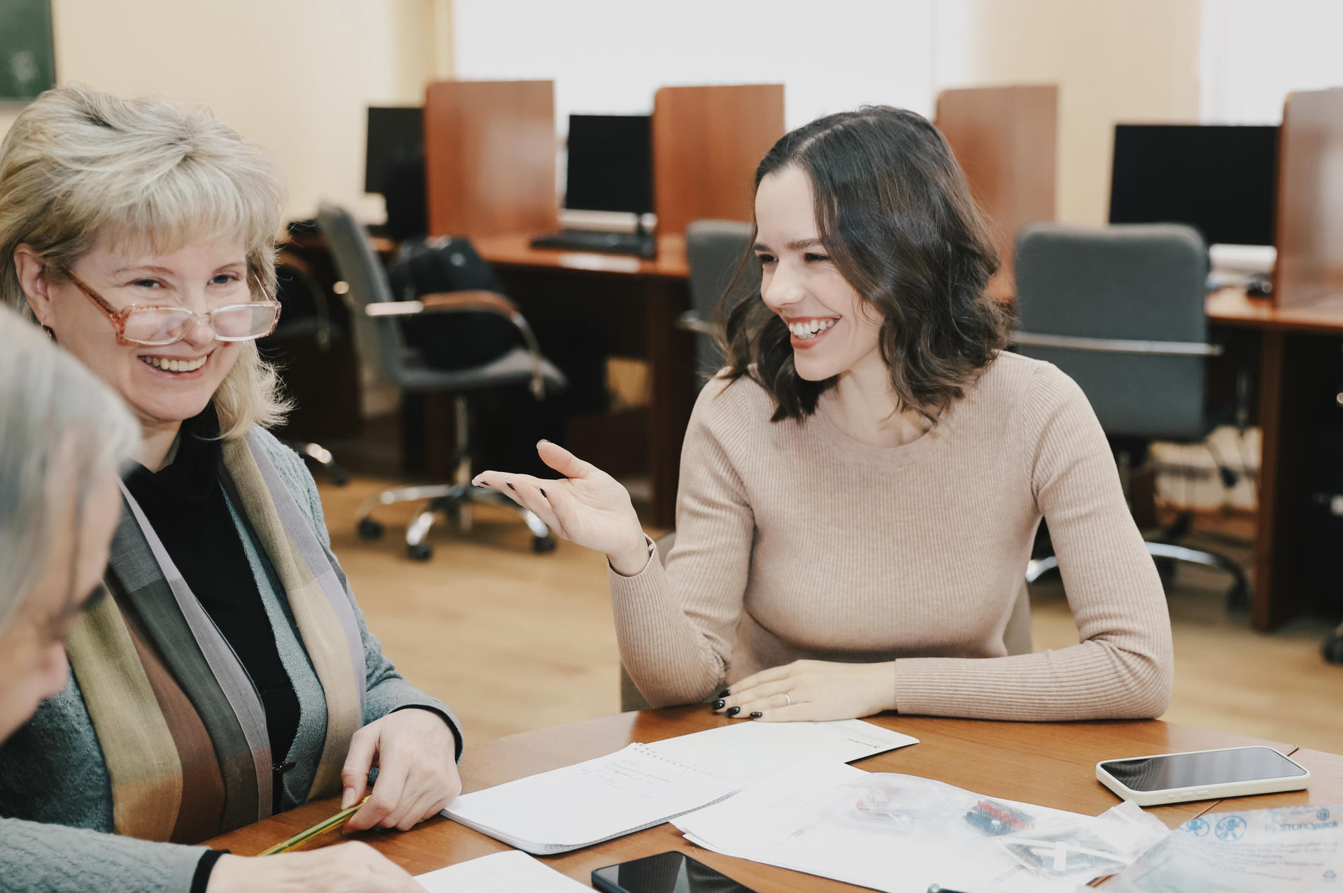 Two women share a light-hearted conversation at a meeting, surrounded by documents on a table.