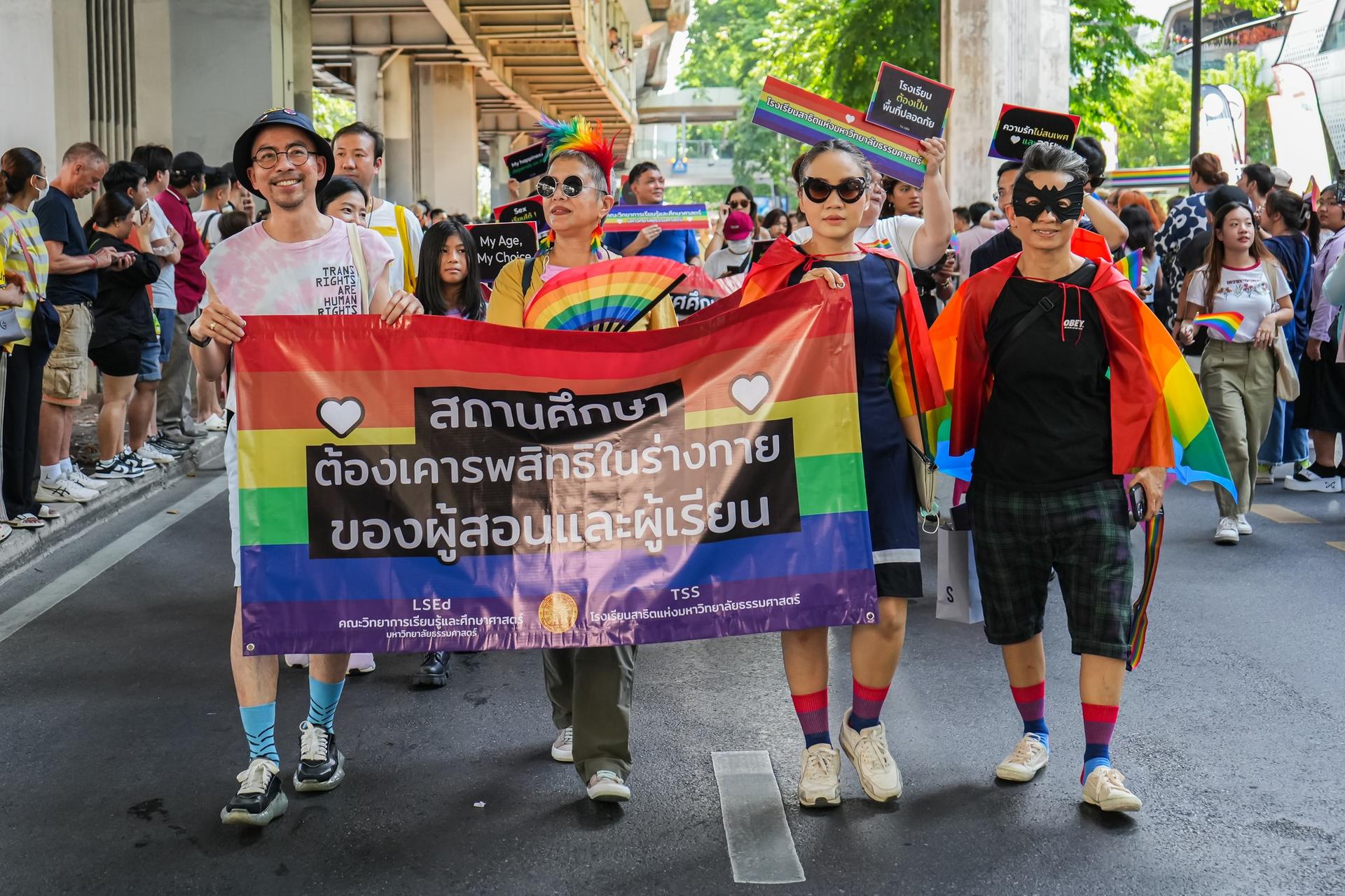 Four people in colorful outfits hold a banner at a pride parade, surrounded by a festive crowd.
