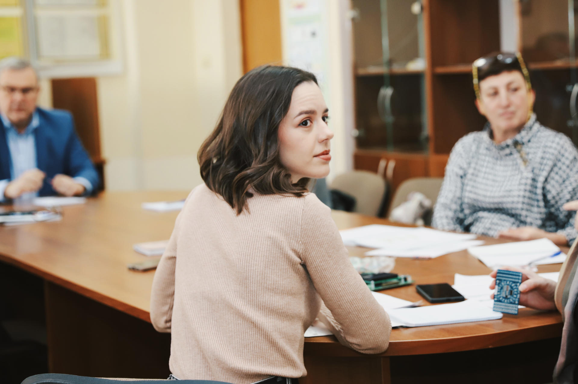 A young woman with dark, wavy hair sits at a table, looking back at others in a meeting.