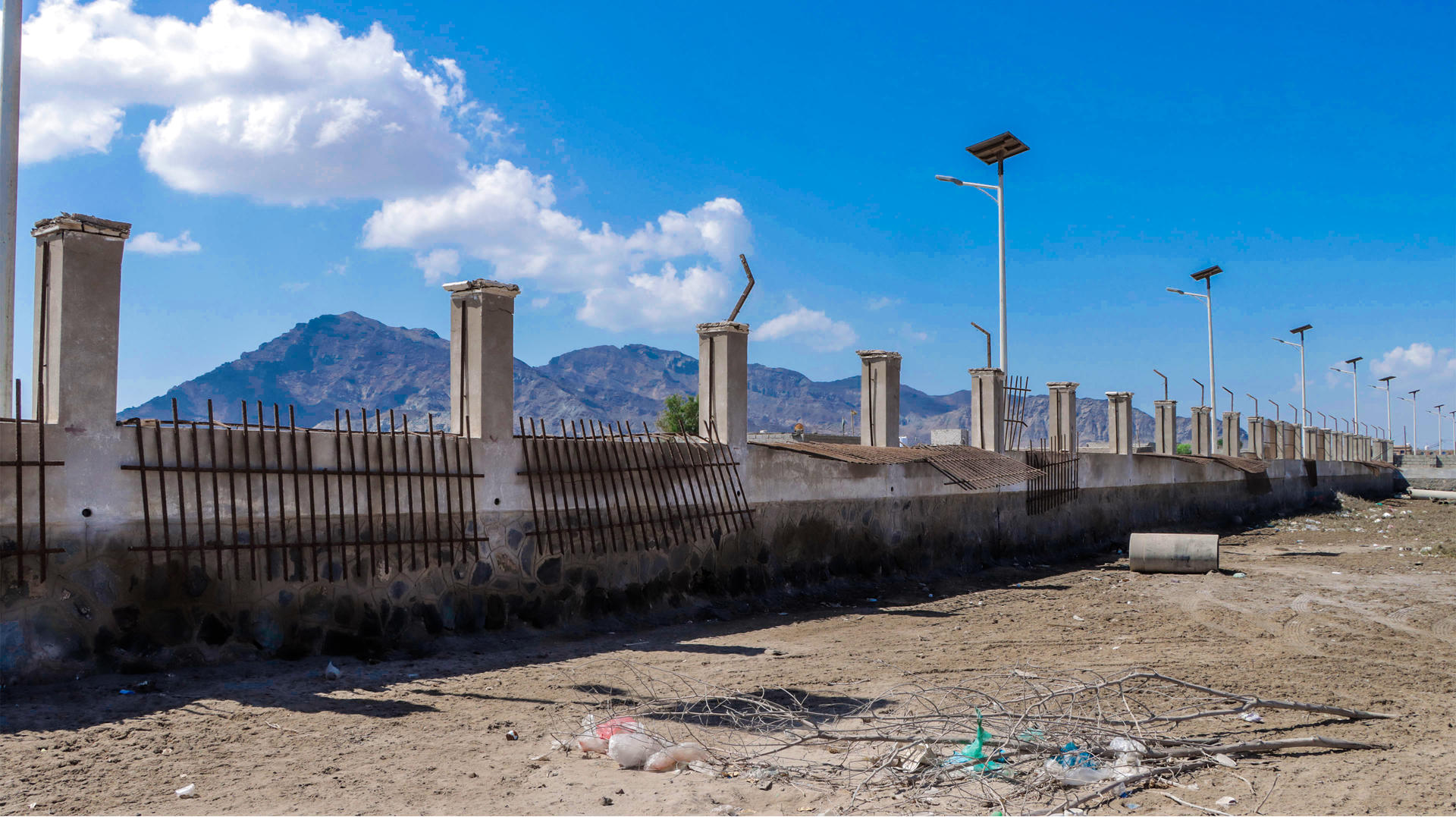 A long, unfinished wall stands against a backdrop of mountains and blue sky.