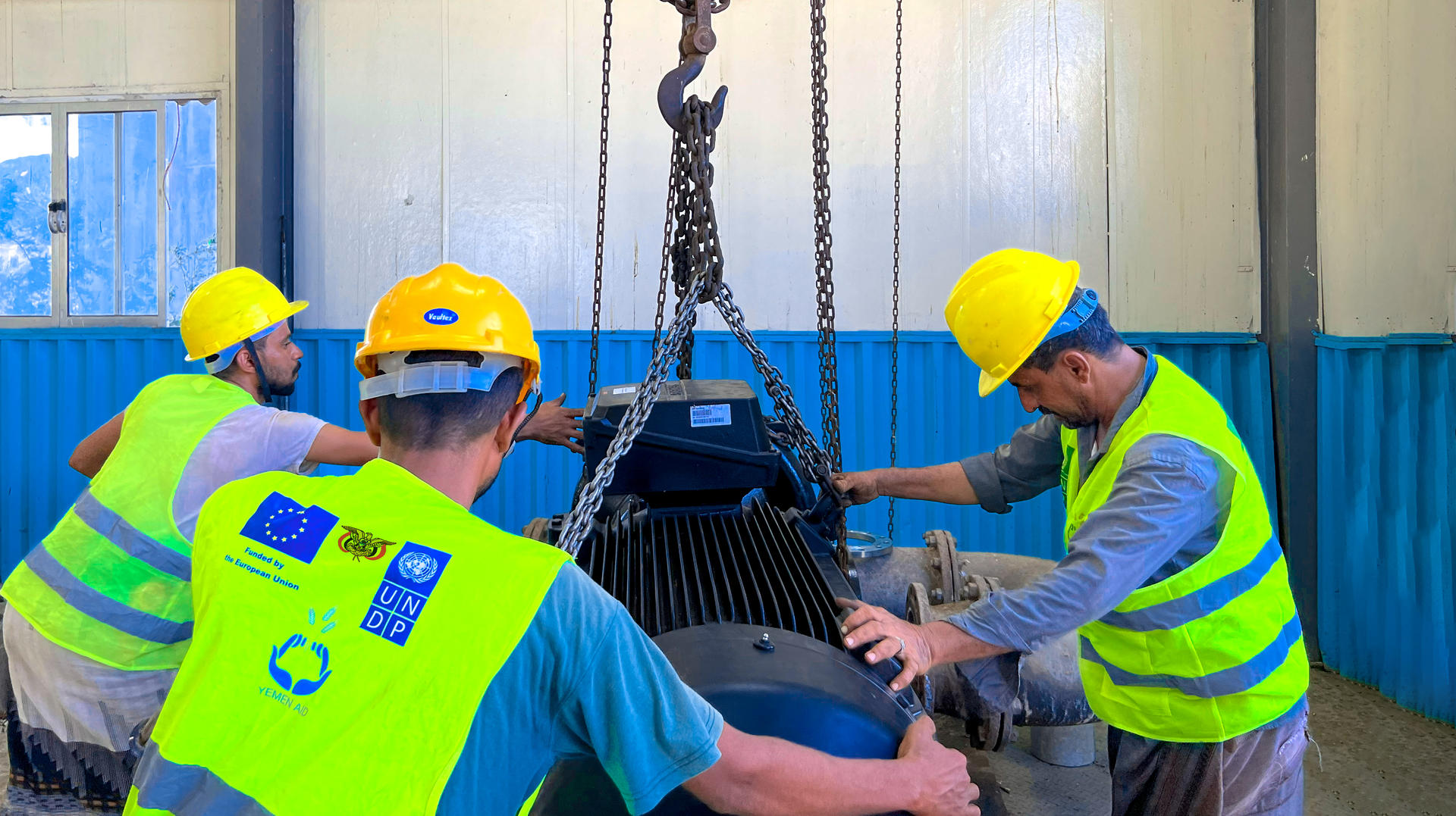 Workers in hard hats and vests carefully maneuver a large machine part in a workshop.