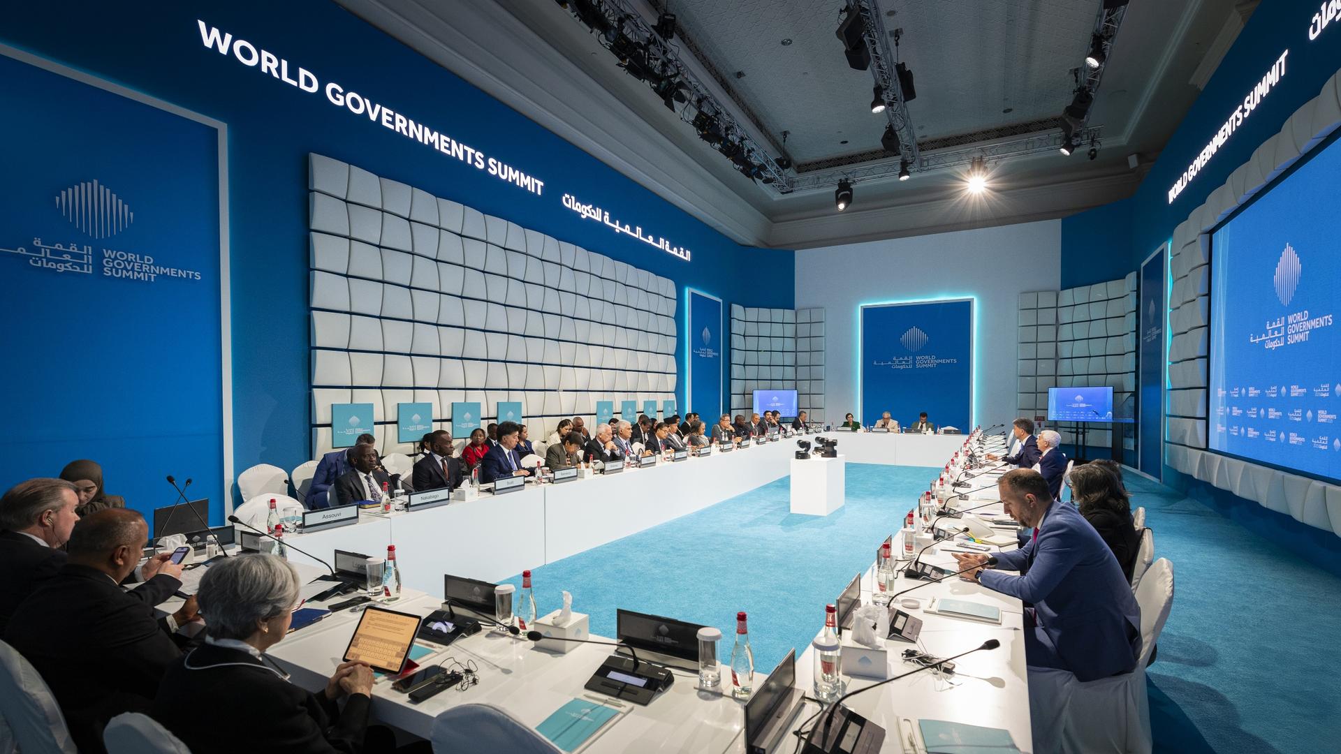 Conference room with attendees seated around a long table under blue decor and screens.