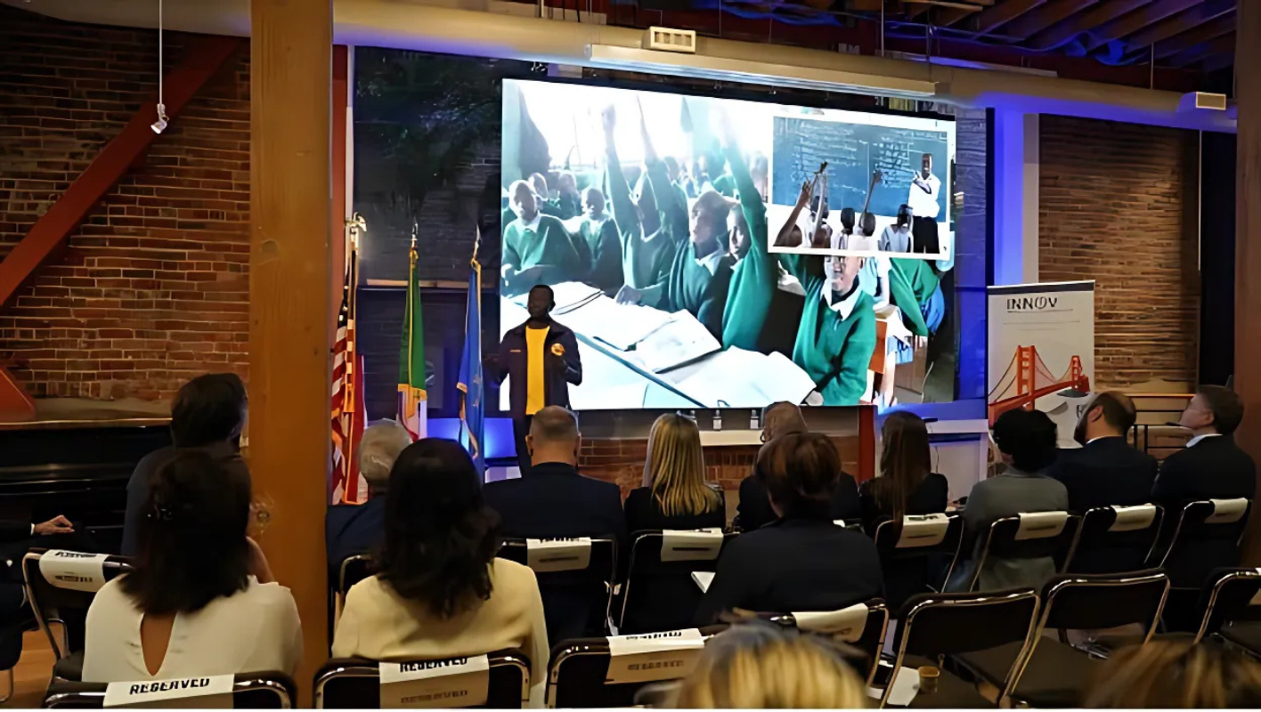 A speaker at a podium addresses an audience in a modern venue with large screens.
