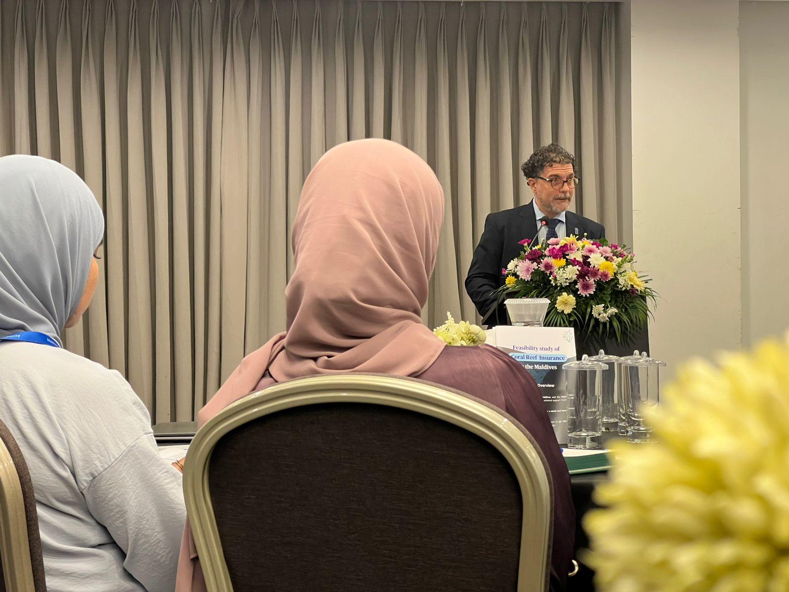 Audience members in headscarves listen to a speaker at a conference with flowers in the foreground.