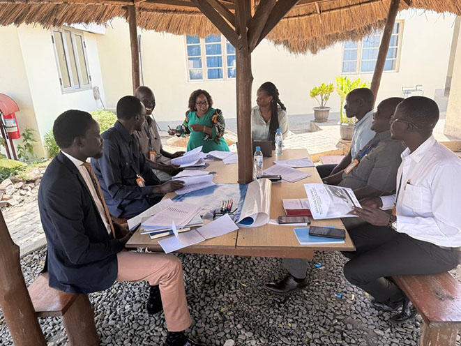 A group of people engaged in a discussion around a table outdoors under a thatched roof.