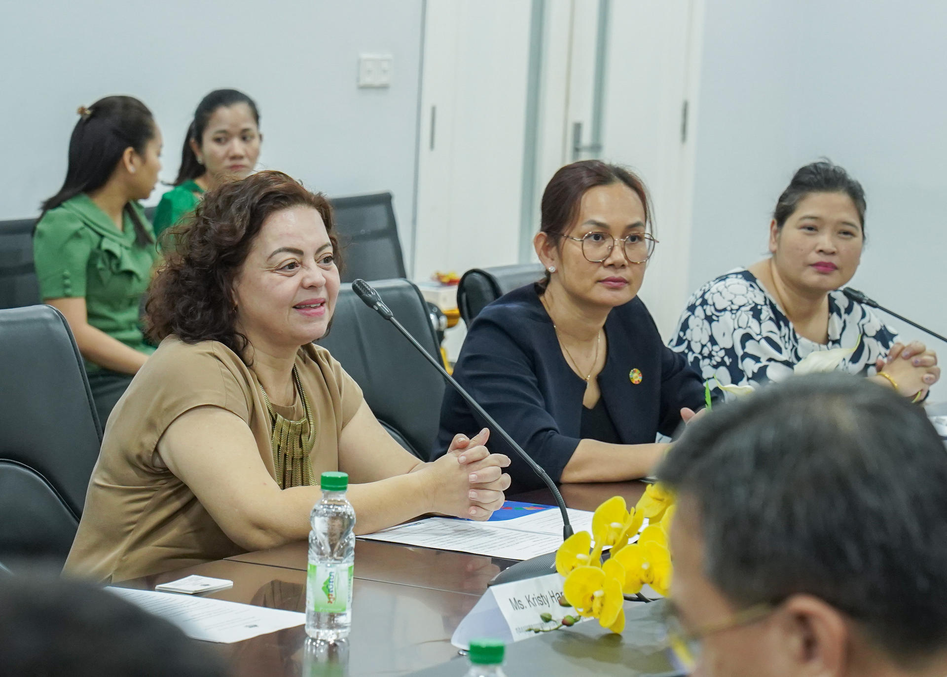 Three women sit at a conference table, engaged in discussion; one smiles warmly.