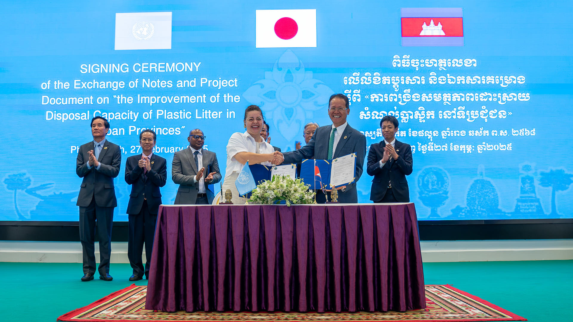 Officials shaking hands over a signed agreement at a ceremonial event with flags in the background.