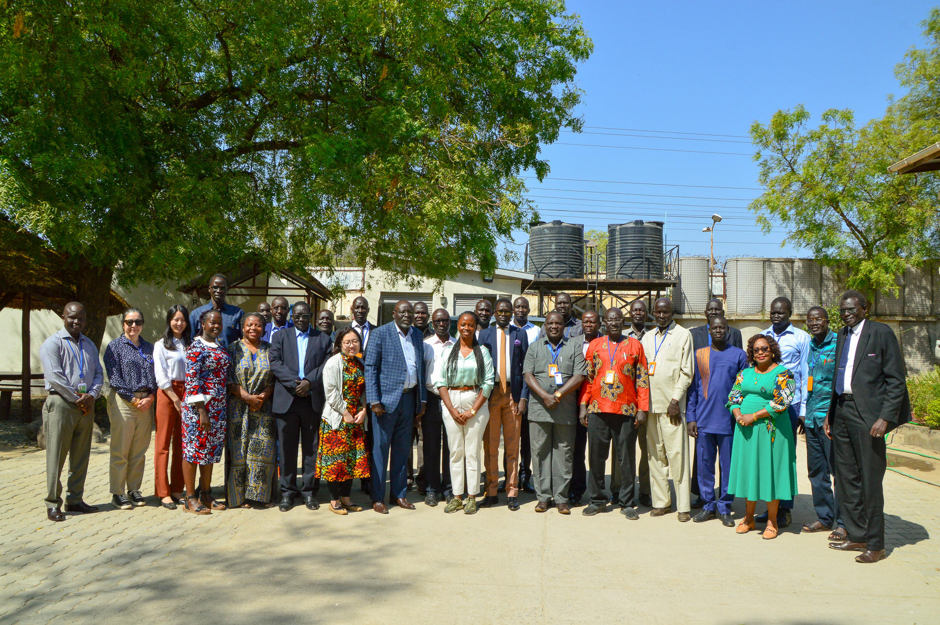 Group of diverse individuals posing outdoors, with a clear blue sky and greenery in the background.