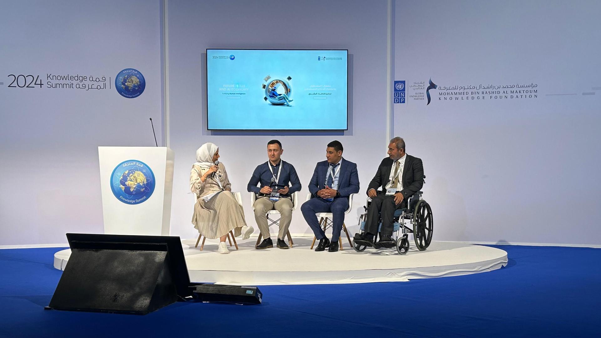 Four panelists seated on a stage, discussing in front of a large screen.