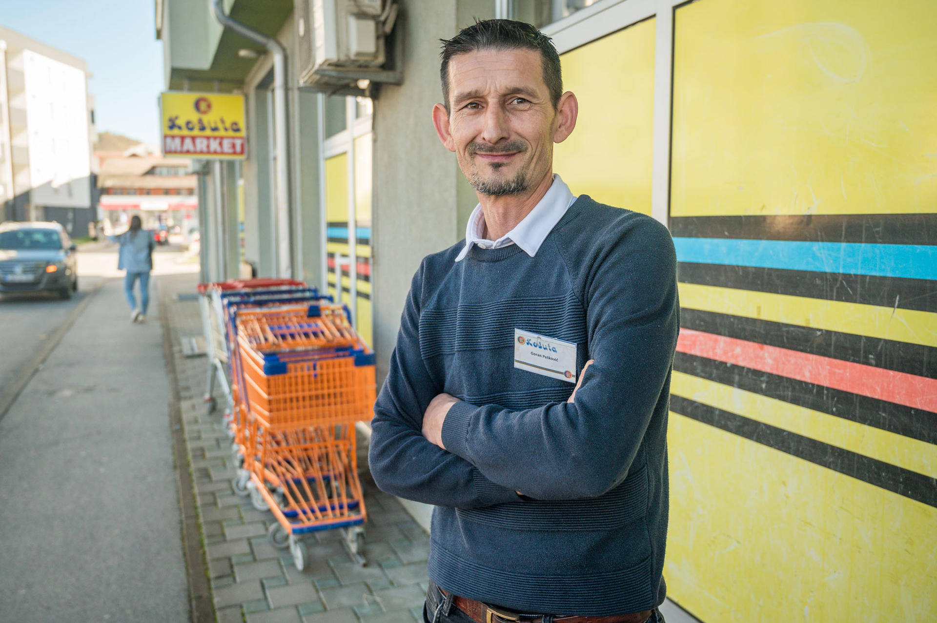 A man stands outside a store with folded arms, smiling, near shopping carts and colorful storefront.