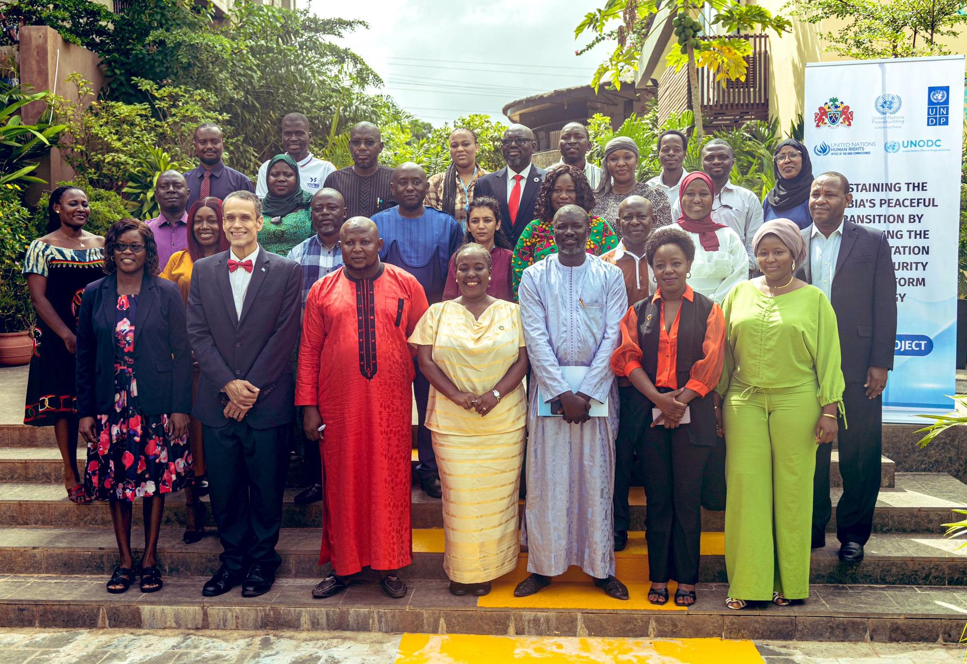 A diverse group of people standing together outdoors, smiling, in formal and traditional clothing.